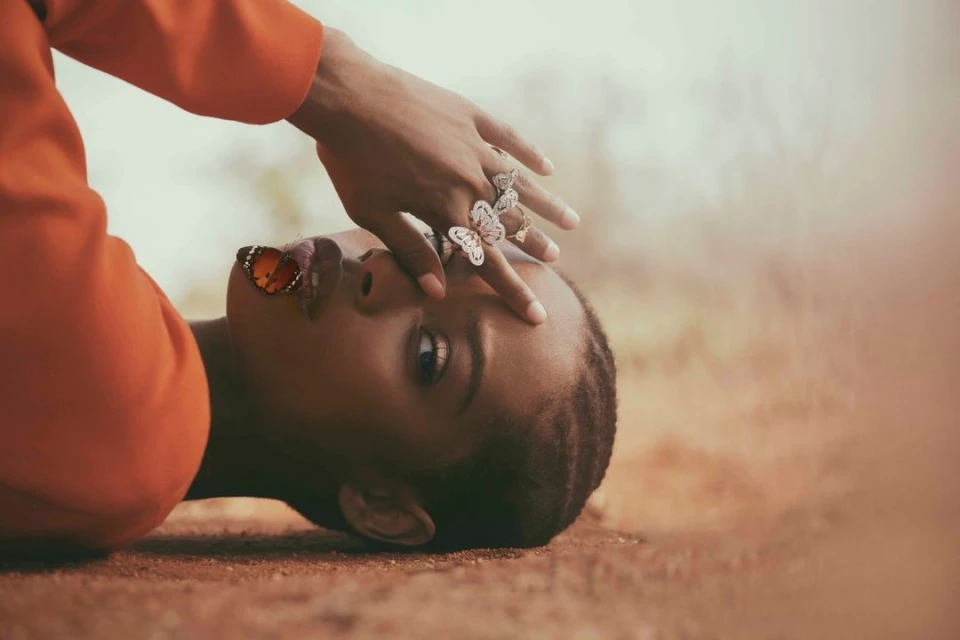 Portrait of a black woman lying down with a butterfly on her lip, wearing butterfly rings and an orange jacket in a soft focus outdoor setting.