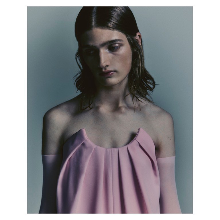 Portrait of a model with long dark hair wearing a pink strapless top against a neutral grey studio background.
