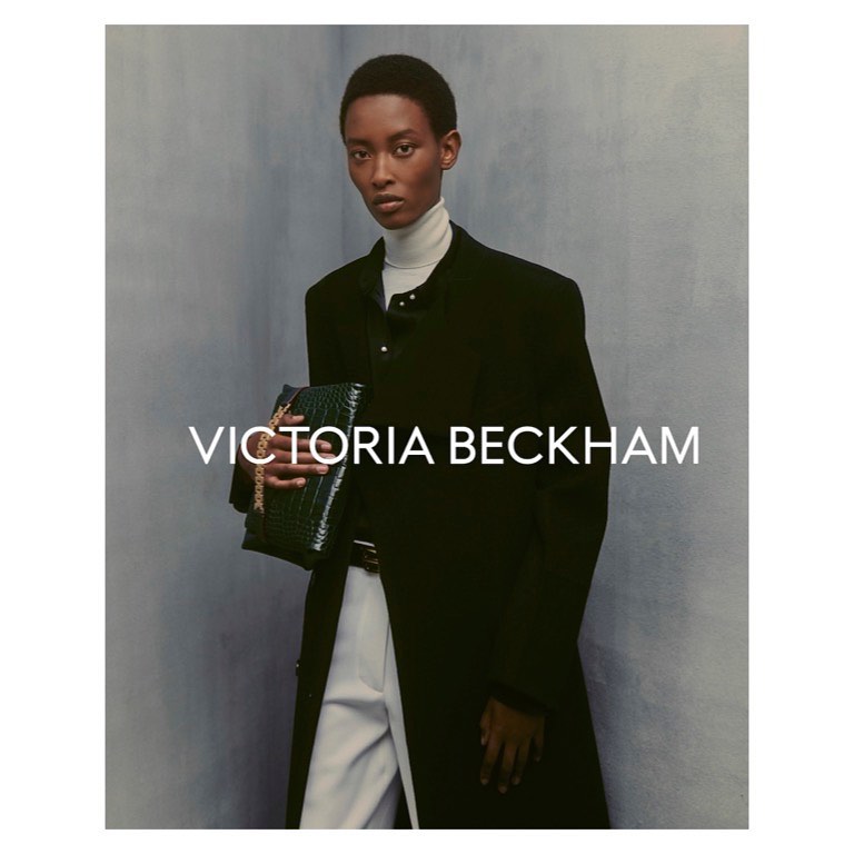 A dark-skinned woman in a black coat holding a green bag. Victoria Beckham logo visible. Studio shot with a muted background.