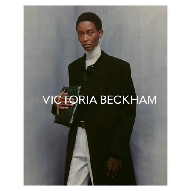 A dark-skinned woman in a black coat holding a green bag. Victoria Beckham logo visible. Studio shot with a muted background.