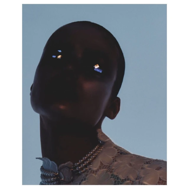 Close-up portrait of a woman with dark skin wearing a white floral lace top and statement jewelry against a light blue background.