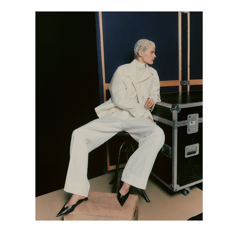 Woman in a cream jacket and white pants sitting on a stool with a black background and a black metal box behind her.