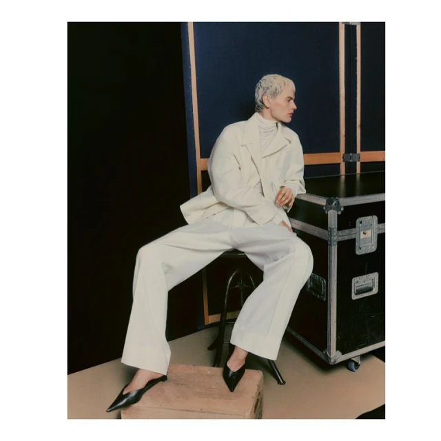 Woman in a cream jacket and white pants sitting on a stool with a black background and a black metal box behind her.