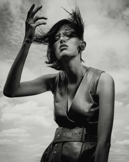 Monochrome portrait of a woman with short dark hair, leather vest, and dramatic hand gesture against a cloudy sky background.