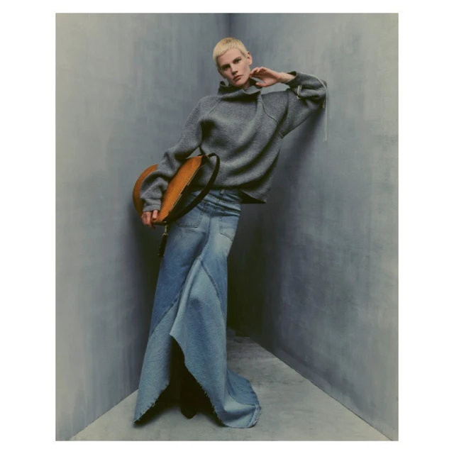 An adult woman with blonde hair poses in a grey sweater and blue denim skirt with a tan leather handbag in a grey corner.