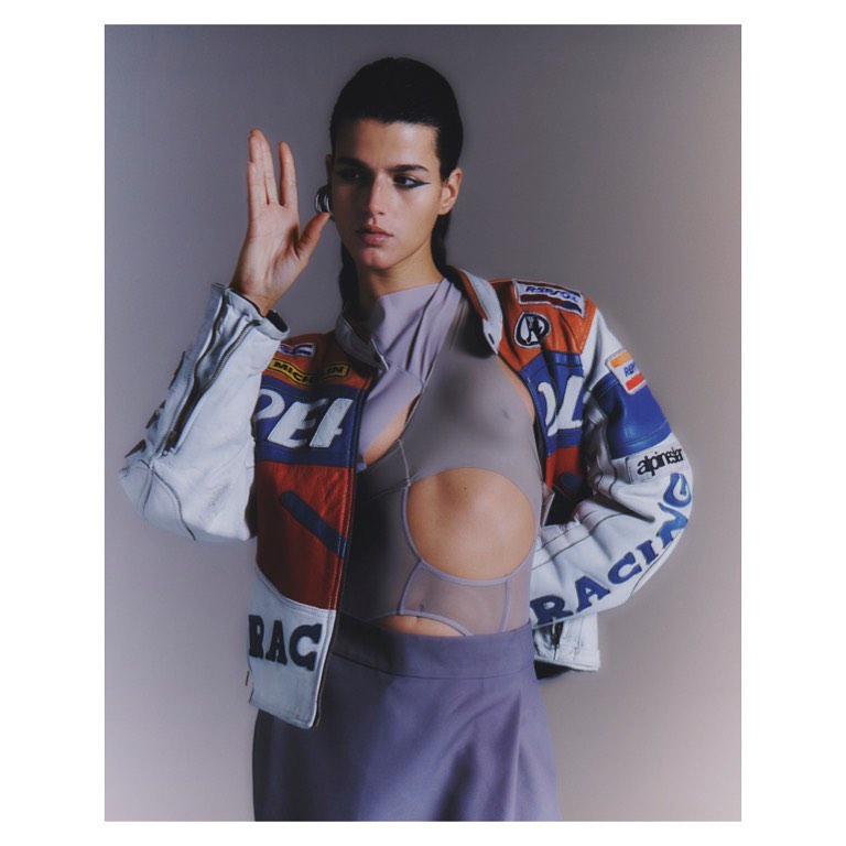 Fashionable woman poses in a cropped cut-out shirt and vintage racing jacket in a studio, showcasing a unique and trendy style.