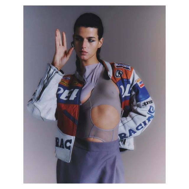 Fashionable woman poses in a cropped cut-out shirt and vintage racing jacket in a studio, showcasing a unique and trendy style.