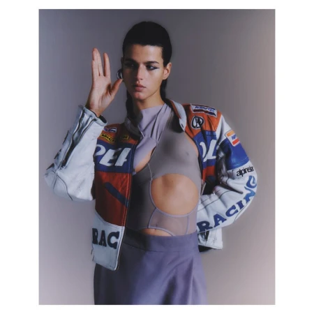 Fashionable woman poses in a cropped cut-out shirt and vintage racing jacket in a studio, showcasing a unique and trendy style.