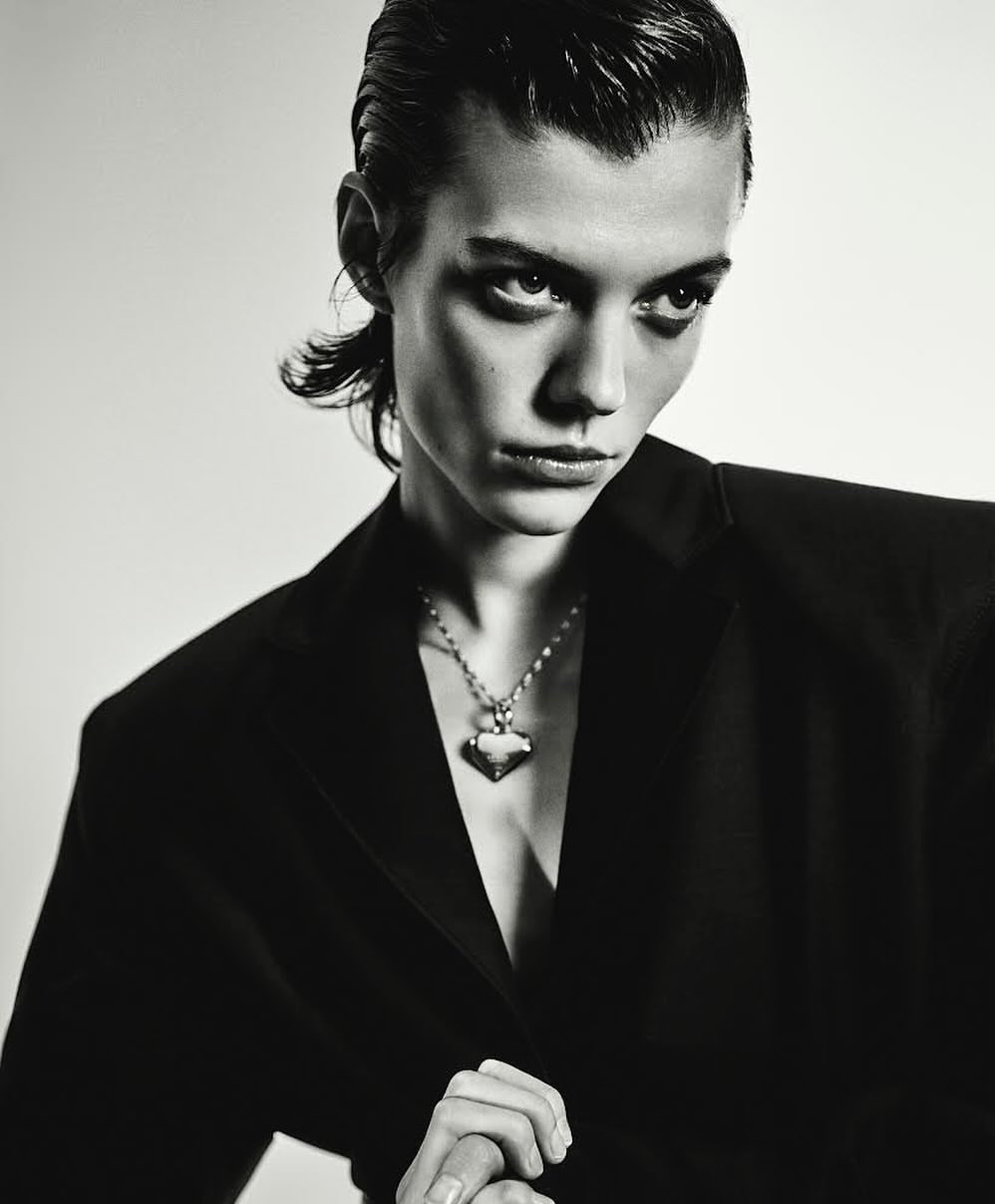 Monochrome portrait of a person wearing a black blazer and silver necklace, set against a stark white background in a studio setting.