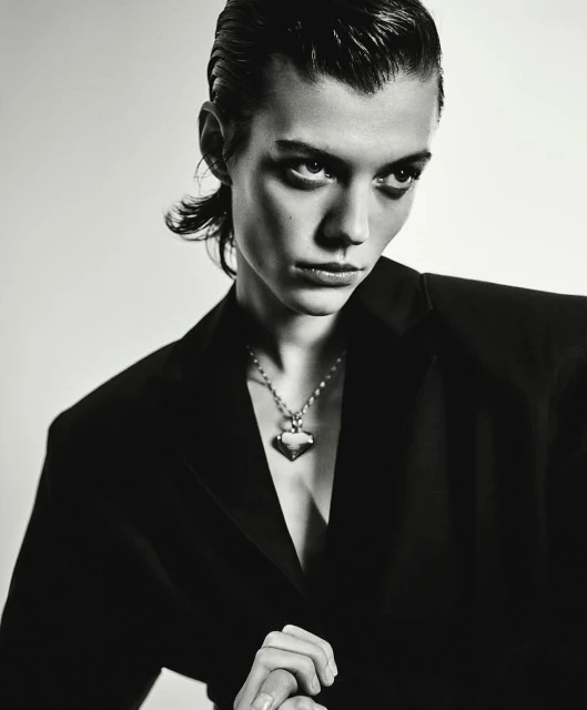 Monochrome portrait of a person wearing a black blazer and silver necklace, set against a stark white background in a studio setting.
