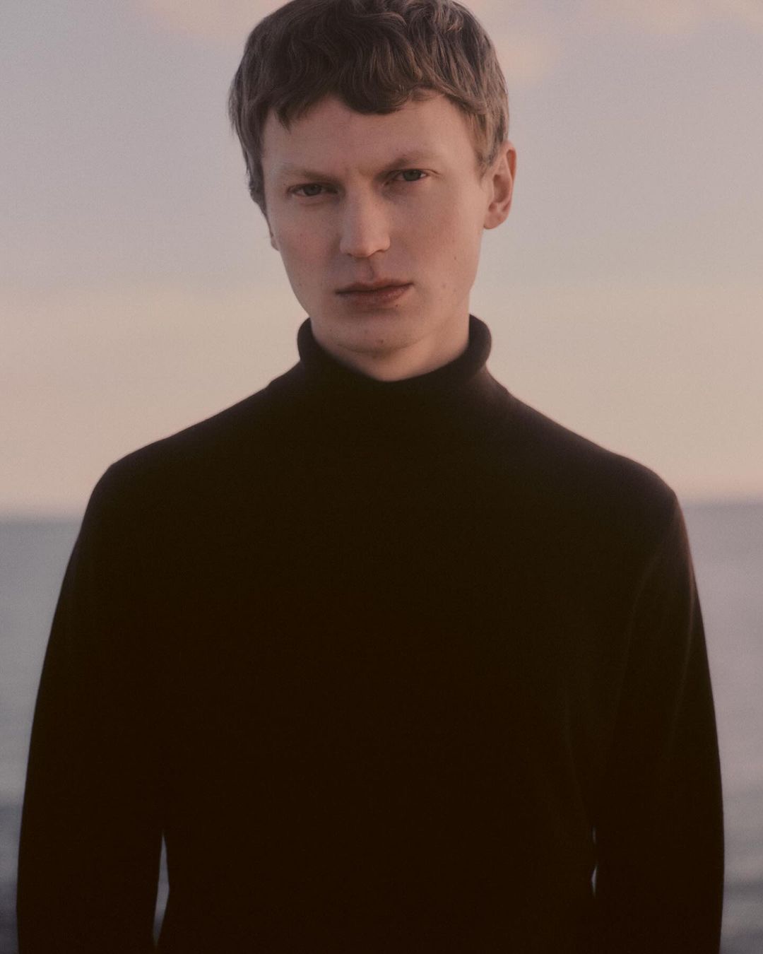 Portrait of a man in his 20s with short dark hair wearing a black turtleneck sweater against an outdoor water background.