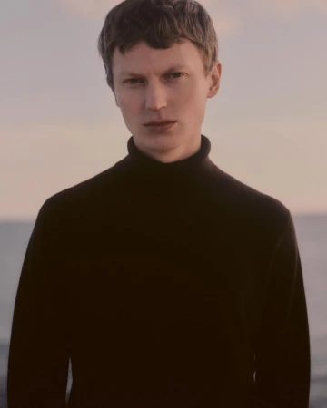 Portrait of a man in his 20s with short dark hair wearing a black turtleneck sweater against an outdoor water background.