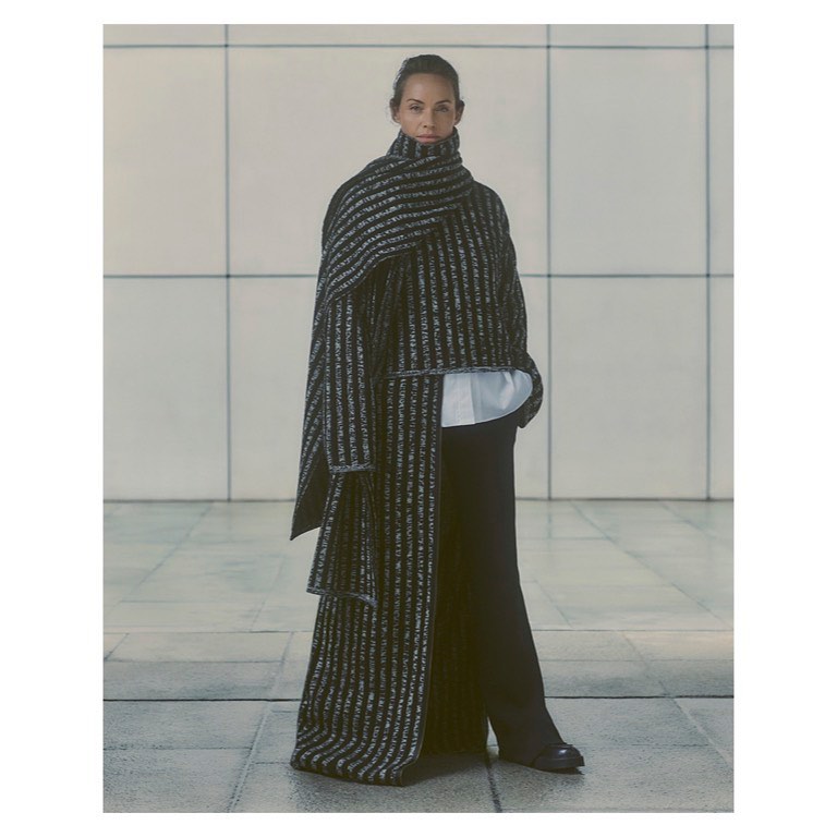 Fashionable adult woman poses in black and silver striped coat with an over-sized wrap, black pants, and black shoes indoors.