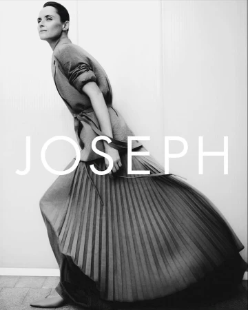 Editorial fashion shot of a woman in a grey dress with pleated skirt posing near a white wall, with the brand name JOSEPH visible.