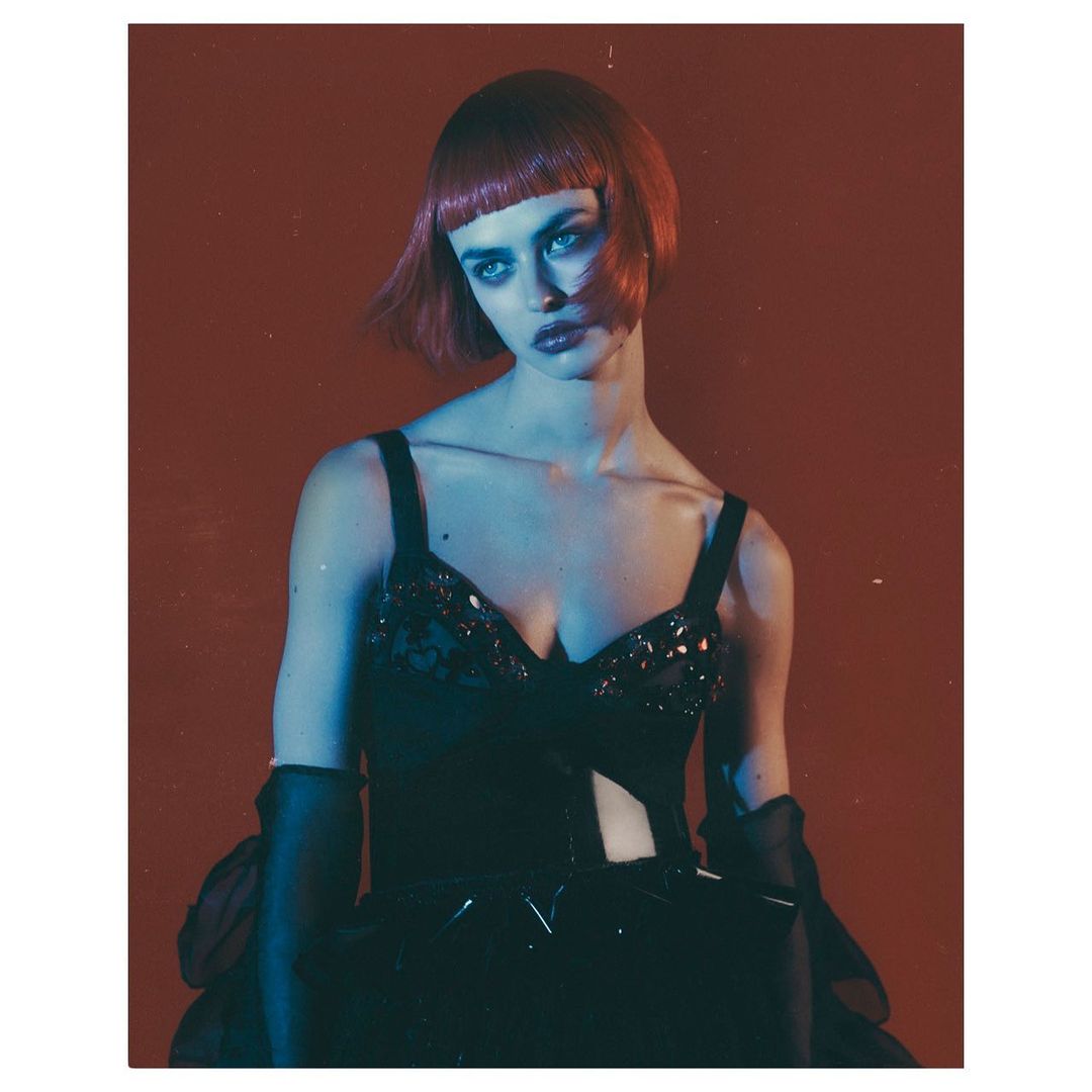 Glamorous adult female in a black embellished dress with dramatic makeup and a red bob hairstyle posing against a vivid red background.