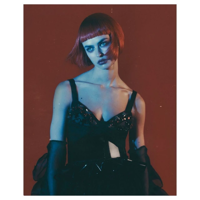 Glamorous adult female in a black embellished dress with dramatic makeup and a red bob hairstyle posing against a vivid red background.