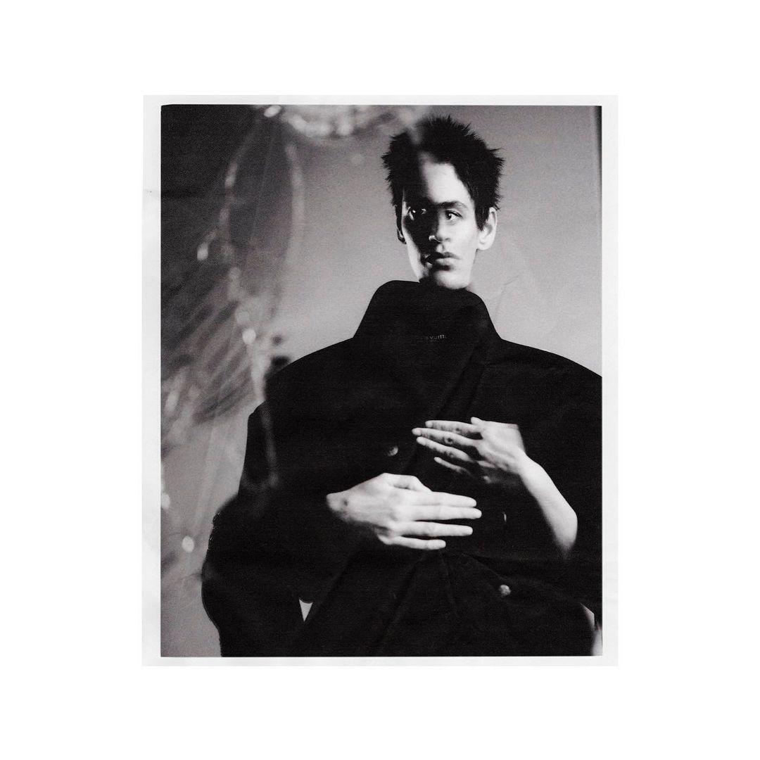 Black and white fashion image of a dark-haired male model in a dark coat with hands placed on his shoulders by another figure.