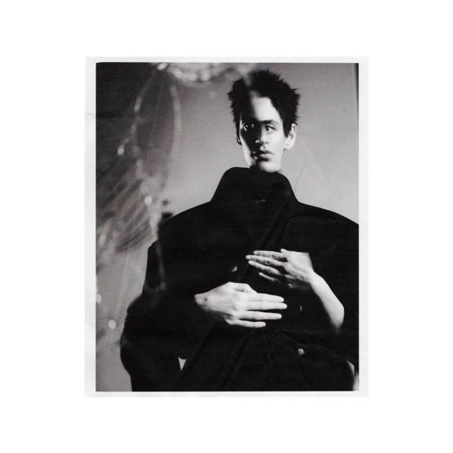 Black and white fashion image of a dark-haired male model in a dark coat with hands placed on his shoulders by another figure.