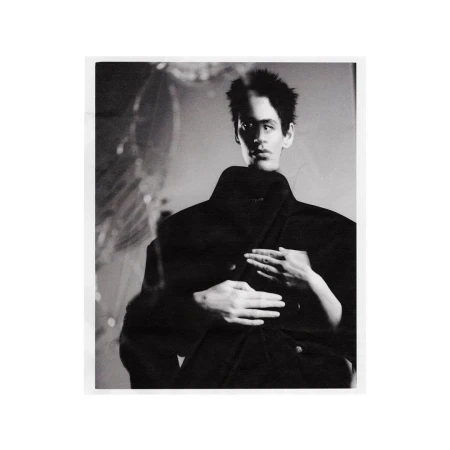 Black and white fashion image of a dark-haired male model in a dark coat with hands placed on his shoulders by another figure.