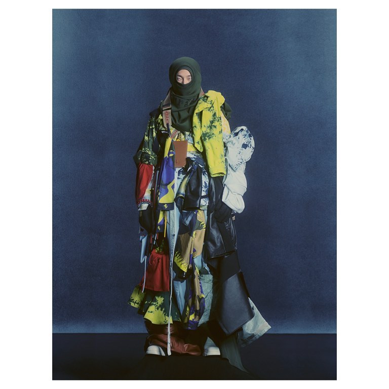 A person wearing a balaclava and colorful, patchwork-style clothing stands against a dark blue background in a fashion-forward studio shot.