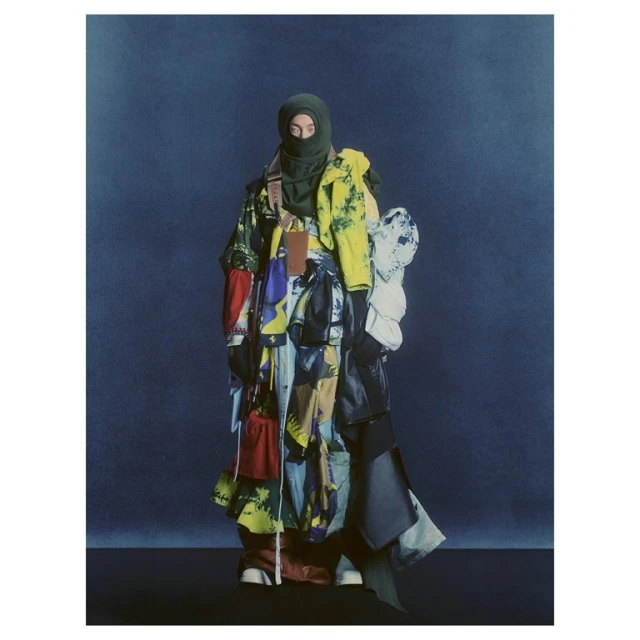 A person wearing a balaclava and colorful, patchwork-style clothing stands against a dark blue background in a fashion-forward studio shot.