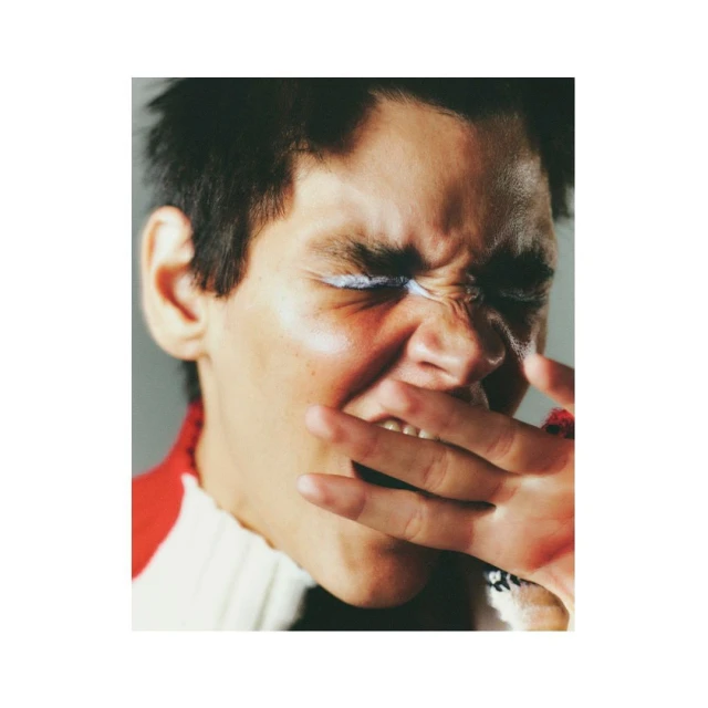 Close-up portrait of an adult with white eyeshadow under closed eyes, dark hair, hand near the mouth, and wearing a red and white top.