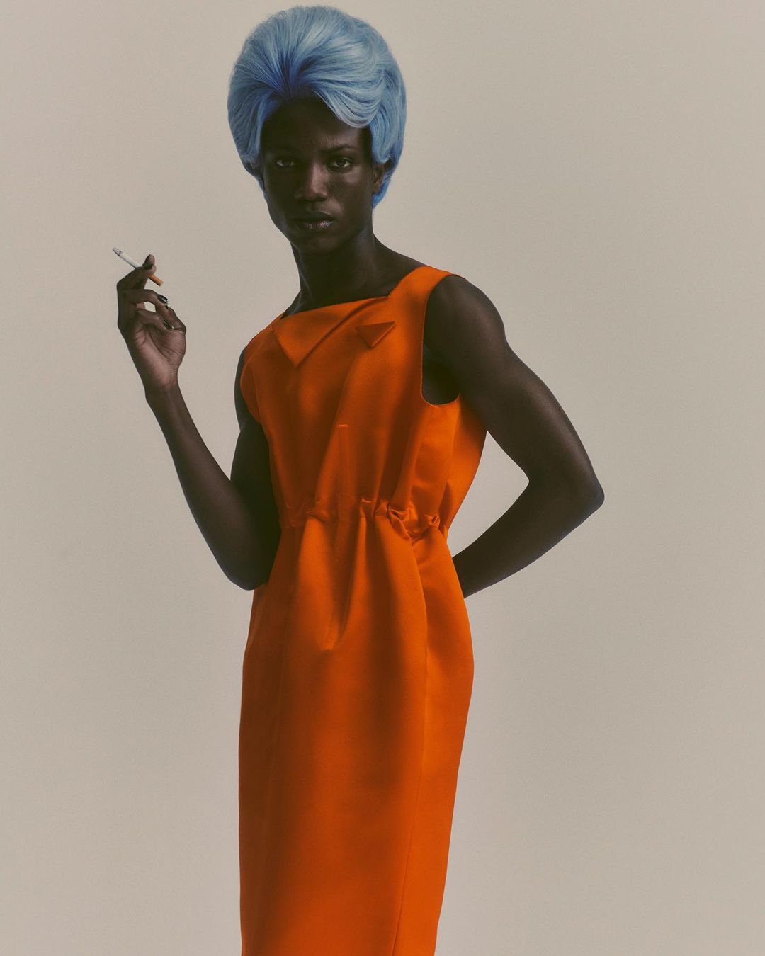 An adult woman with dark skin poses indoors wearing a bright orange dress and a blue wig while holding a cigarette in her hand.
