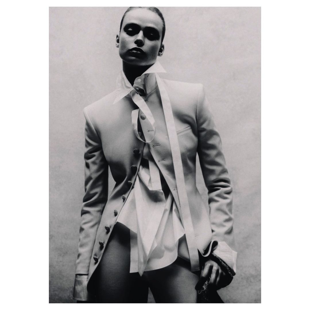 Monochrome studio shot of a young adult fashion model in a blazer and dress shirt, posing with dramatic lighting against a neutral backdrop.