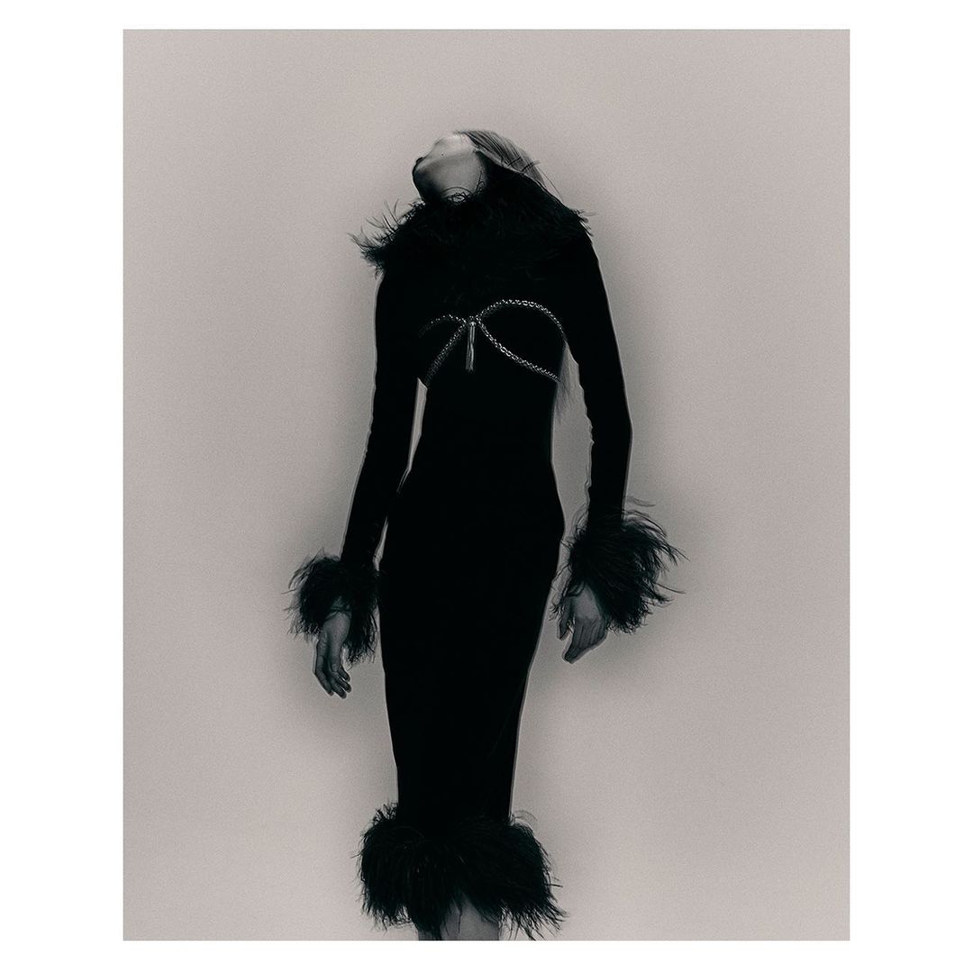 Monochrome fashion shot of a woman in a form-fitting black dress with feather trim, posing with her head tilted back in a studio.