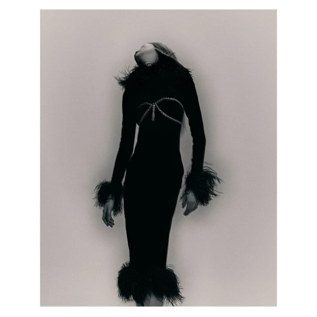 Monochrome fashion shot of a woman in a form-fitting black dress with feather trim, posing with her head tilted back in a studio.