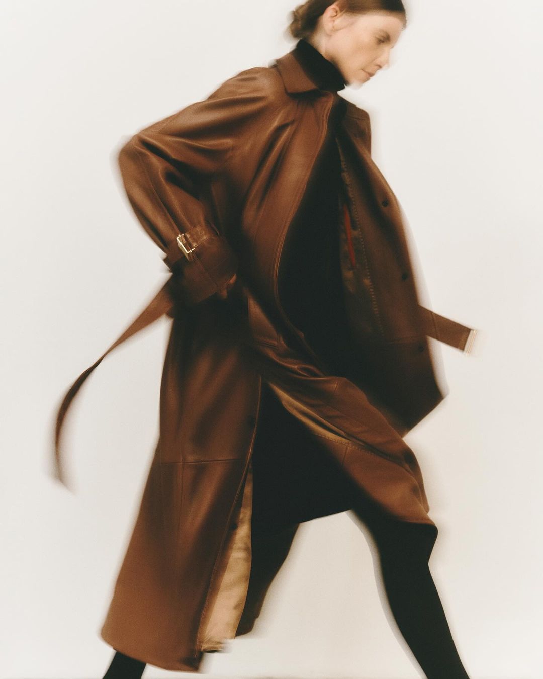 Model in motion wearing a long brown leather trench coat against a plain light background