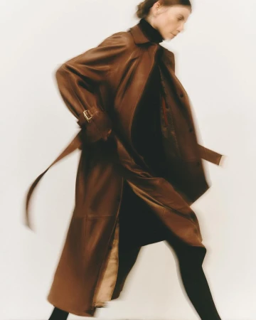 Model in motion wearing a long brown leather trench coat against a plain light background