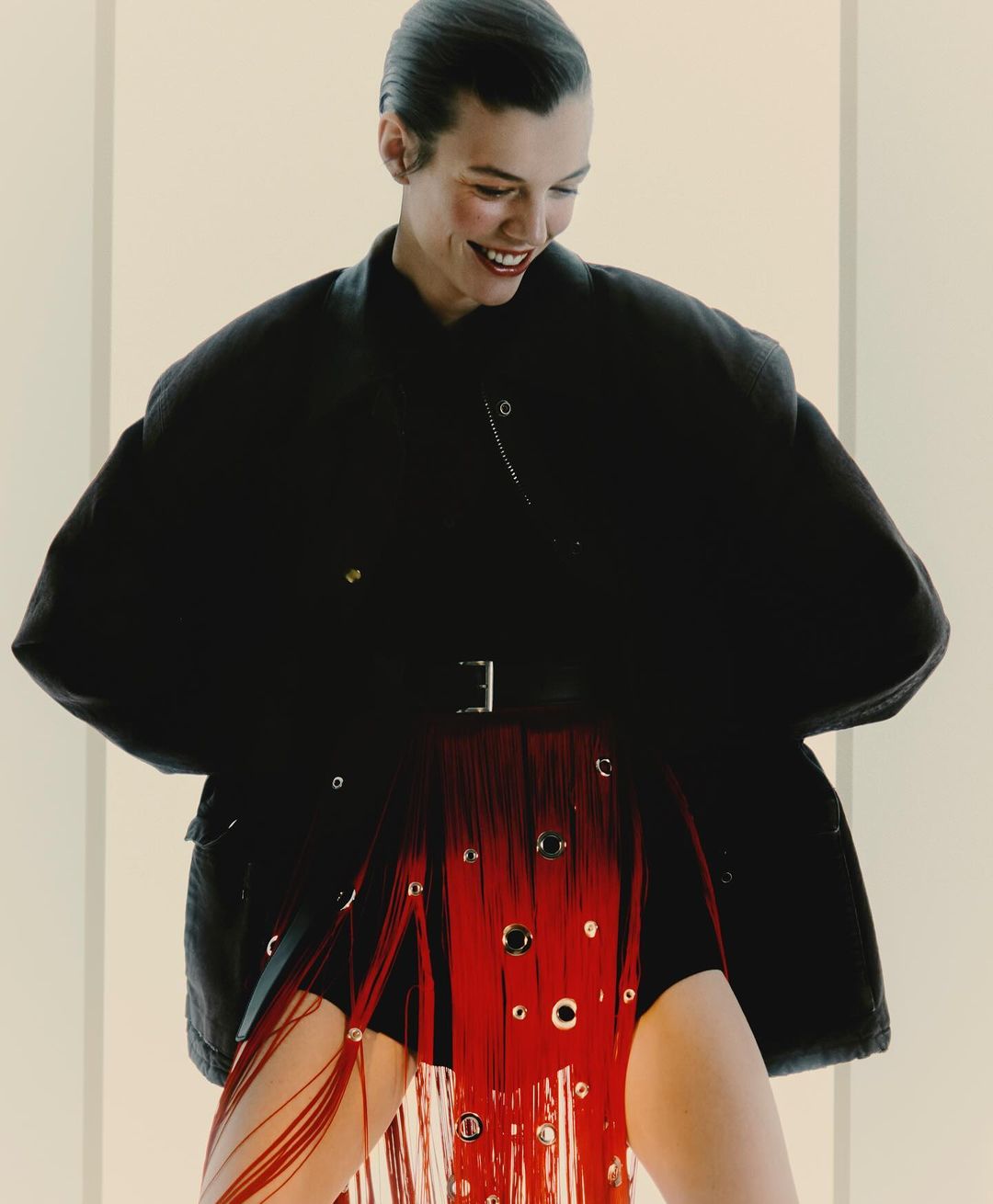 A woman smiles in a studio wearing a black jacket, shorts, and a red fringe skirt with metal eyelets, in a fashion shot.