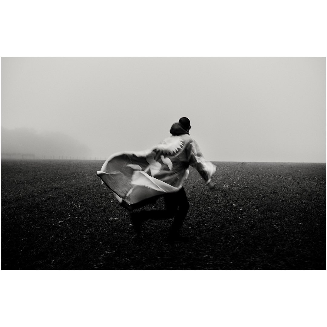 Person runs through a grassy field on a foggy day wearing a patterned cape or blanket, captured in black and white.