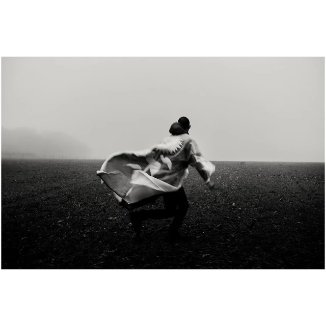 Person runs through a grassy field on a foggy day wearing a patterned cape or blanket, captured in black and white.