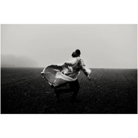 Person runs through a grassy field on a foggy day wearing a patterned cape or blanket, captured in black and white.