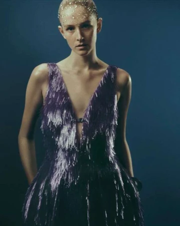 Model in a deep purple fringe dress with bead embellishments on her forehead against a dark blue background.