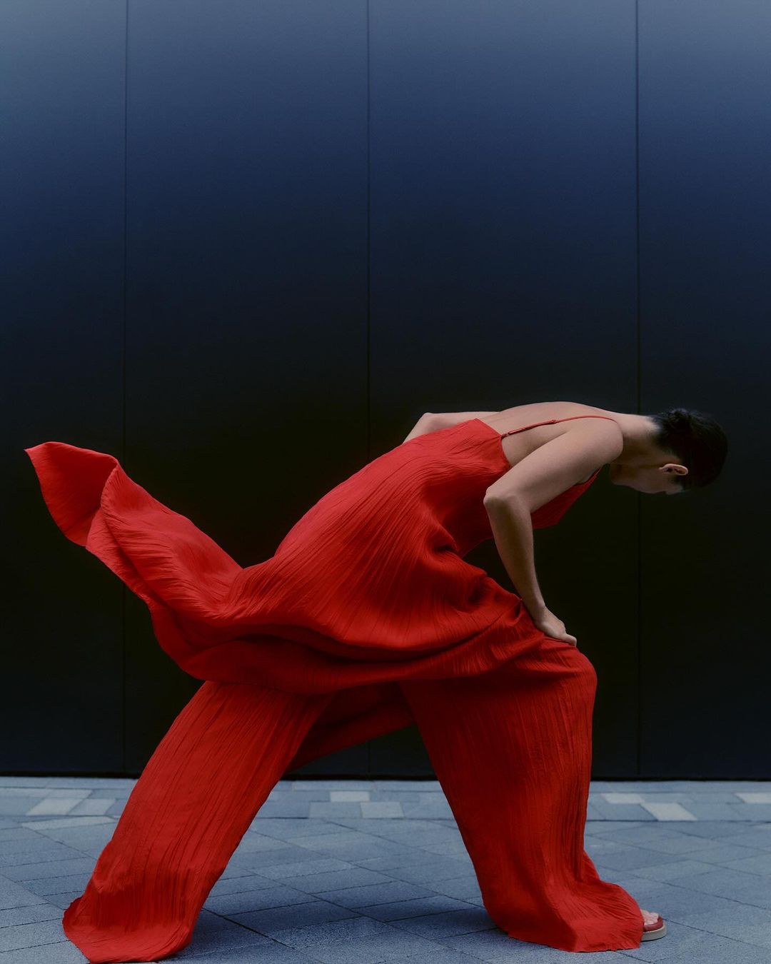 Model in a bright red textured jumpsuit leaning forward against a dark panelled wall outdoors in London.