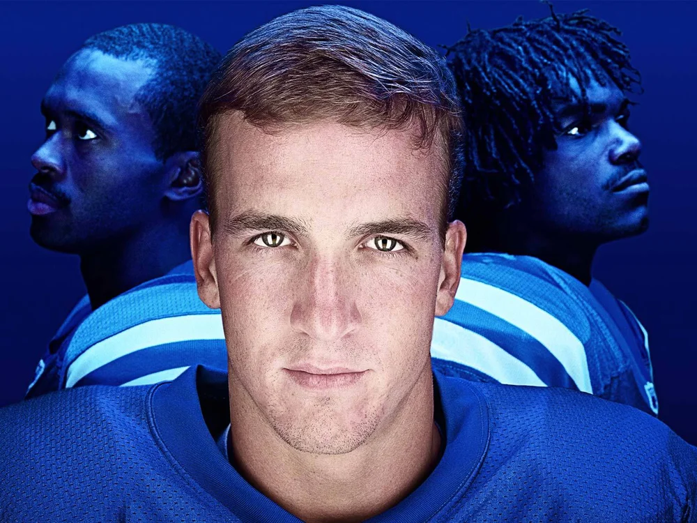 Close-up of three American football players in blue uniforms against a dark blue backdrop, featuring central figure with light skin.
