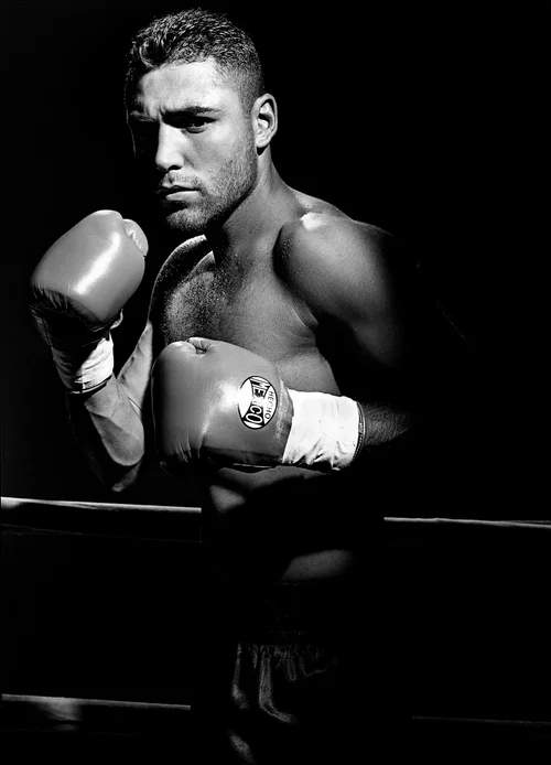 Monochrome portrait of a muscular adult male boxer with boxing gloves in a boxing ring, dramatic lighting, and intense expression.