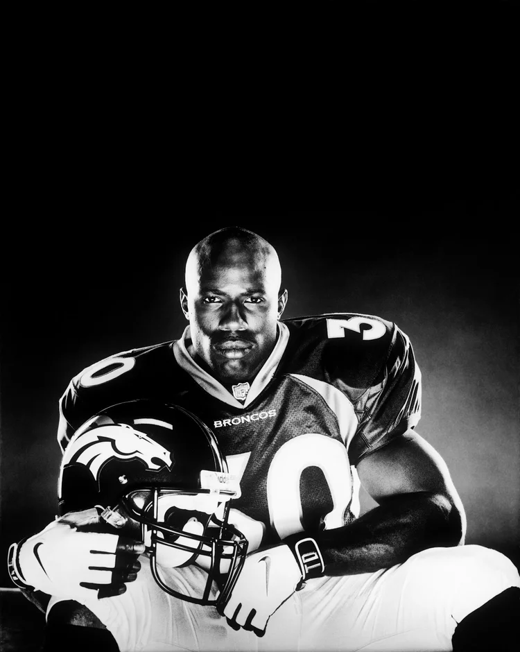 Portrait of a muscular football player in uniform holding a helmet, set against a stark black backdrop in black and white.