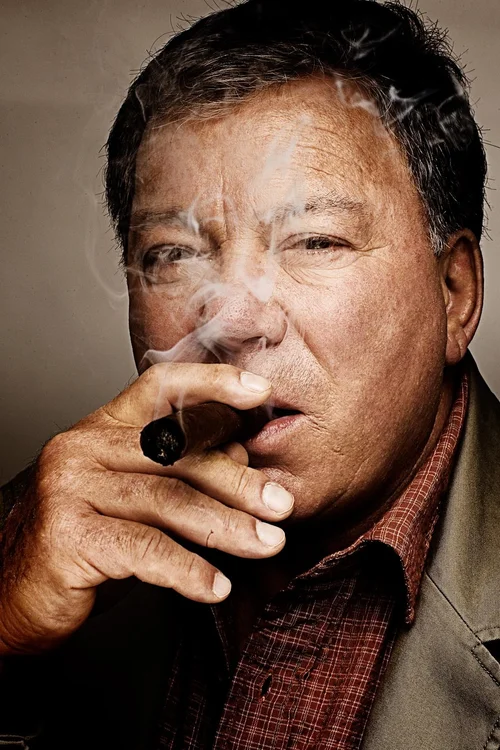 Close-up portrait of William Shatner with a serious expression, smoking a cigar. He wears a red plaid shirt and a tan jacket.
