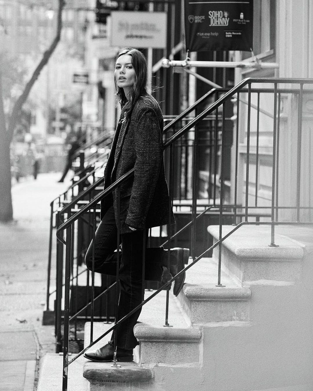 Woman in a tweed blazer and black jeans stands on grey outdoor steps with black iron railings in a black-and-white lifestyle shot.