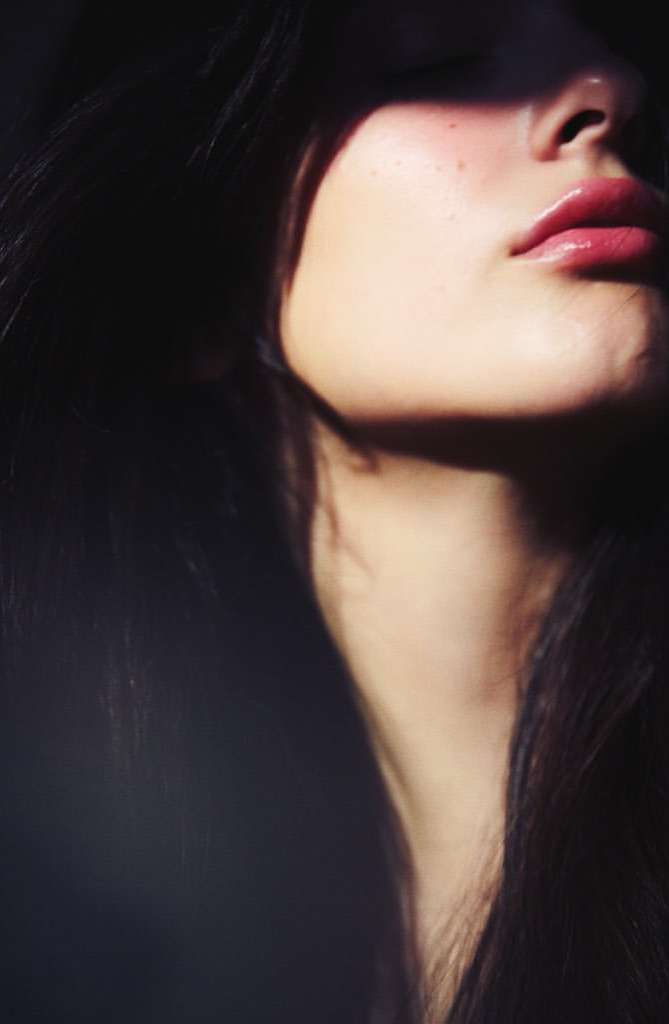Close-up beauty portrait of a young adult woman with dark hair and pink lipstick, partially lit with light and shadow.
