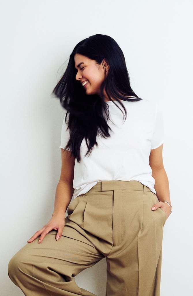 Smiling brunette woman in a white tee and khaki wide-leg pants poses indoors against a white wall, showcasing a casual fashion style.