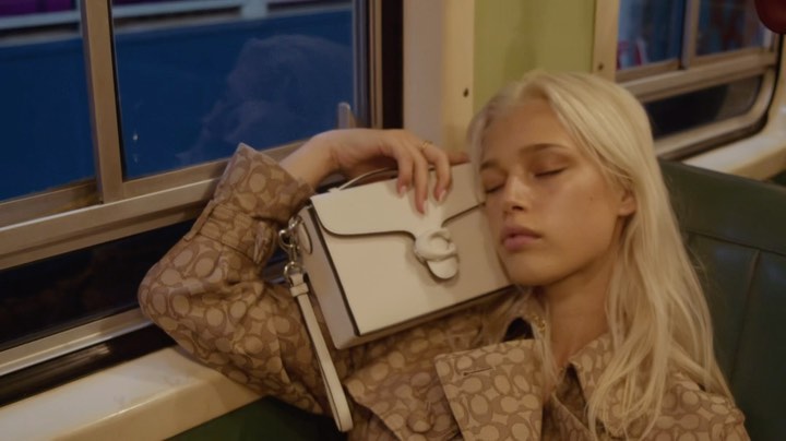 Woman with white-blond hair rests, holding a white purse with silver chain detail in train-like setting, wearing patterned tan coat.