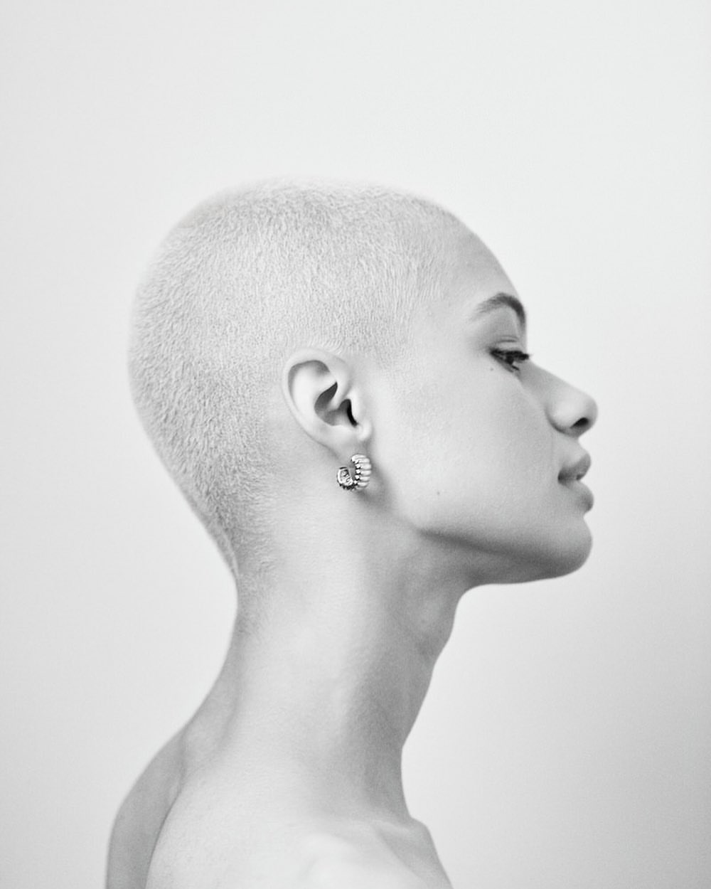 Monochrome profile shot of an adult female with a shaved head wearing hoop earrings against a plain white background in a photo studio.