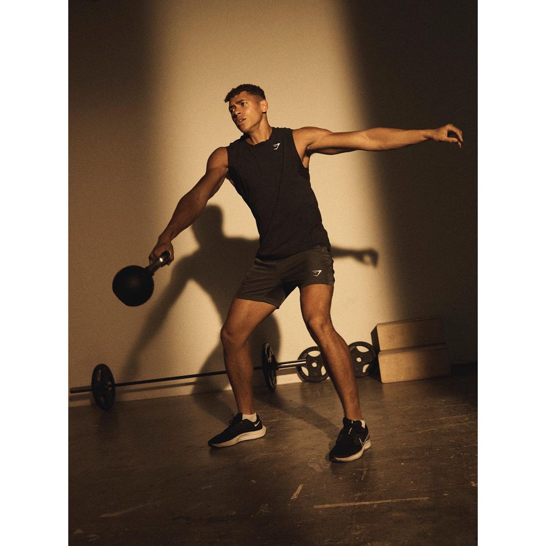 Athletic man in tank top and shorts holding kettlebell in gym, weight training with a barbell in the background in studio lighting.