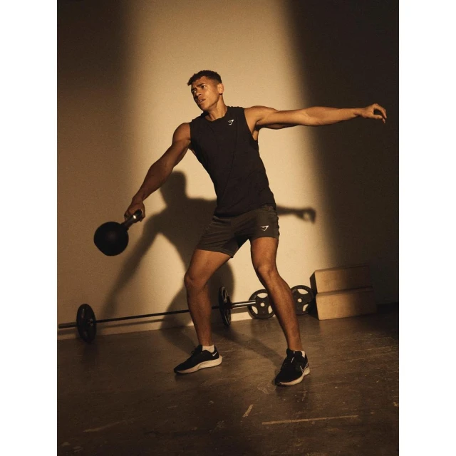 Athletic man in tank top and shorts holding kettlebell in gym, weight training with a barbell in the background in studio lighting.