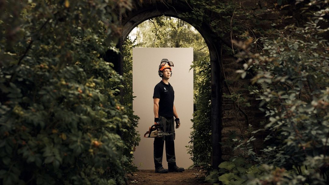 Man stands holding a chainsaw in front of a garden archway, wearing a protective helmet with ear protection, in an outdoor setting.